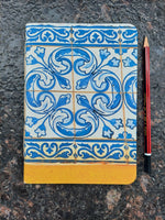 An A5 size notebook with a blue and white Portuguese tile pattern cover and a yellow-orange spine, shown on a dark textured stone surface with a red pencil below it.
