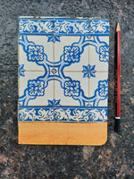 A hardcover notebook with a blue and white floral tile pattern cover and a tan leather spine, shown on a dark speckled stone surface with a red pencil below it.