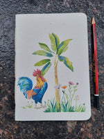 A small rectangular sketchbook or card with a colorful painted rooster perched next to a banana tree, placed on a dark mottled stone surface with a red and black pencil below it.