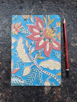 A rectangular notebook or journal with a vibrant blue and pink floral block-print pattern, shown on a dark textured stone surface with a red pencil below it.