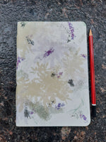 An open notebook with a floral watercolor cover design in muted beige, purple, and green tones, placed on a dark textured surface with a red pencil underneath.